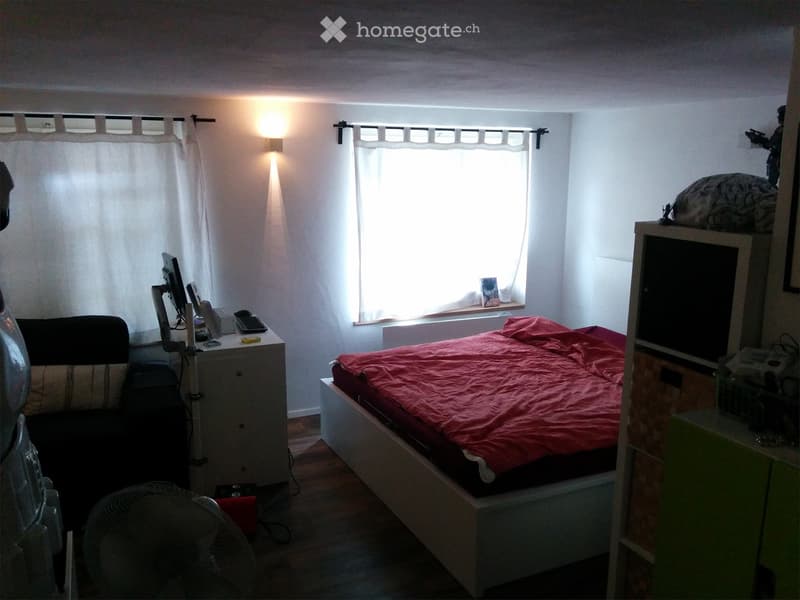 A small bedroom with a bed covered in a red blanket, a fan on the floor, a chair, a screen, and a table with various items on it. The room has wooden flooring and white walls with curtains on the windows.