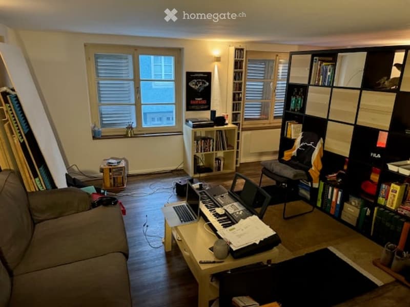 A cozy living room with a sofa, a small table holding a laptop and a keyboard, a bookshelf, a chair, and a piano. The room has wooden flooring and large windows with blinds.