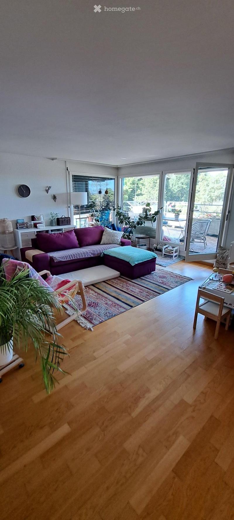 Modern living room with wooden flooring, large windows, and a balcony. Features a purple sectional sofa, coffee table, side table, and plants.