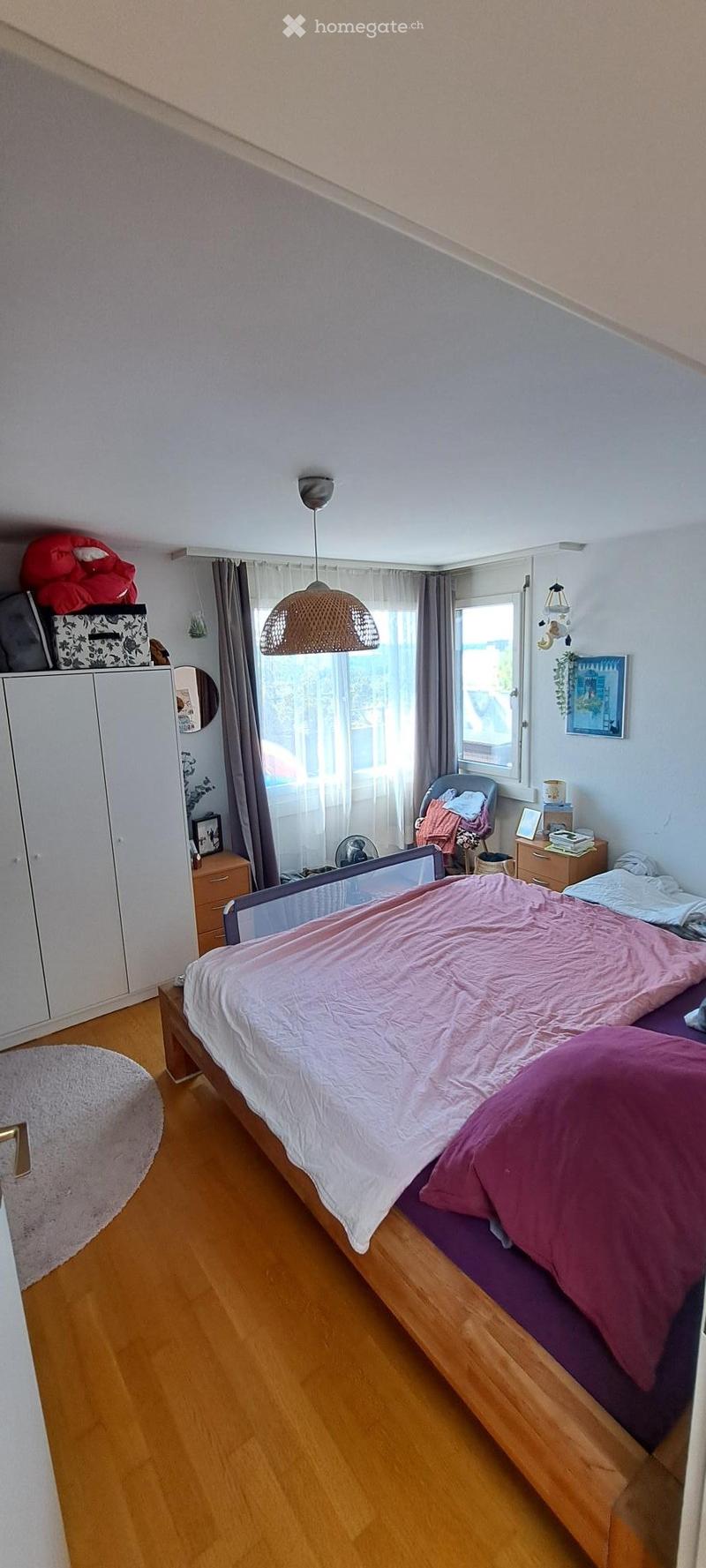 Bedroom with wooden floor, large window with curtains, bed with pink bedding, wardrobe, nightstand, chair, and fan.