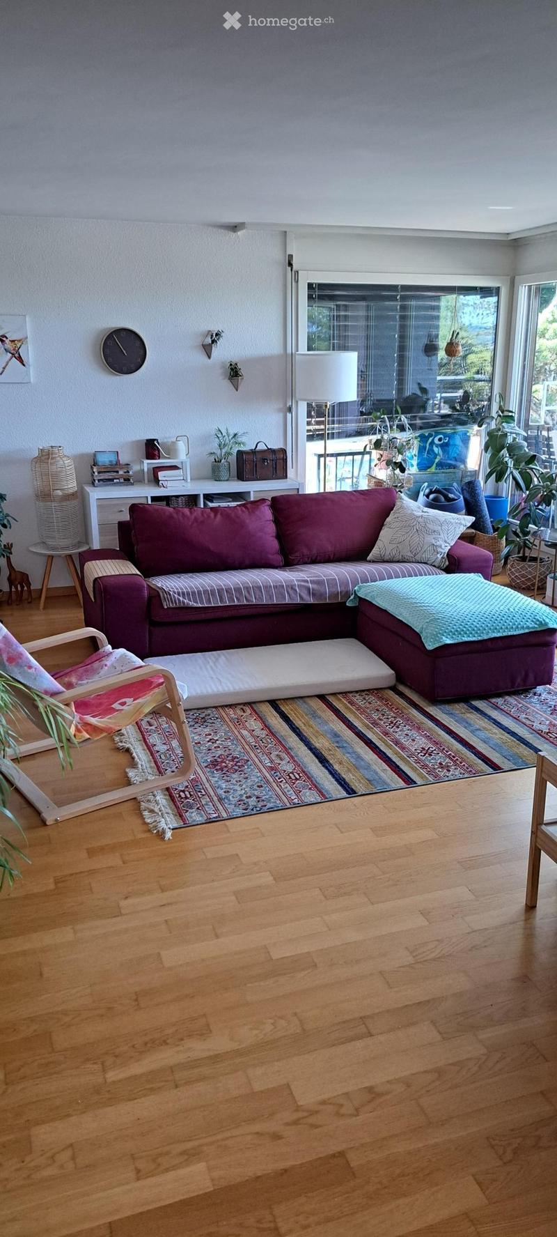 Modern living room with wooden flooring, a purple sectional sofa, an ottoman, a wooden chair, a glass coffee table, a patterned rug, and decorative items including a vase, books, and a clock on a white shelf.