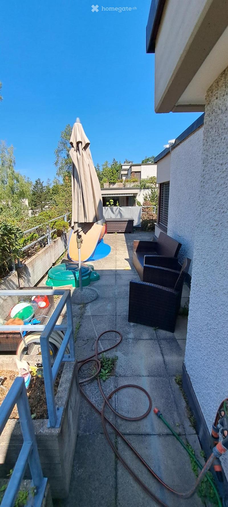 Balcony with concrete floor, umbrella, chairs, table, and garden hose.