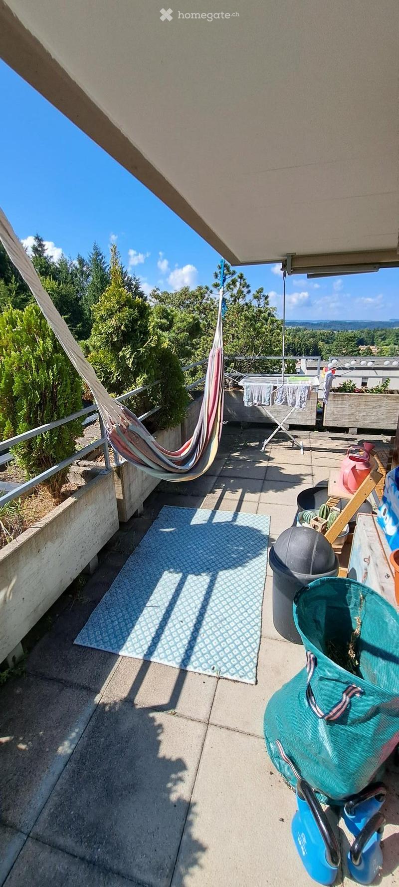 Balcony with a hammock, rug, trash bin, and garden tools, offering a scenic view of trees and hills.