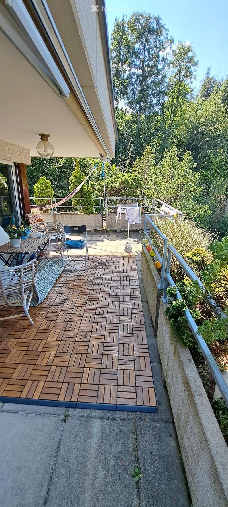Balcony with wooden decking, metal railing, and outdoor furniture including a table, chairs, and a hammock.