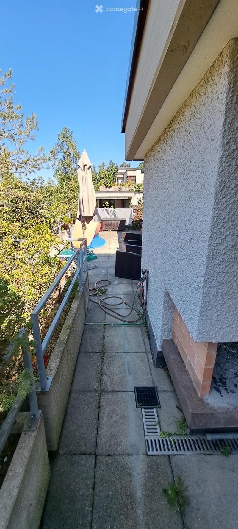 Narrow concrete balcony with metal railing, outdoor furniture, and a view of a garden and neighboring building.