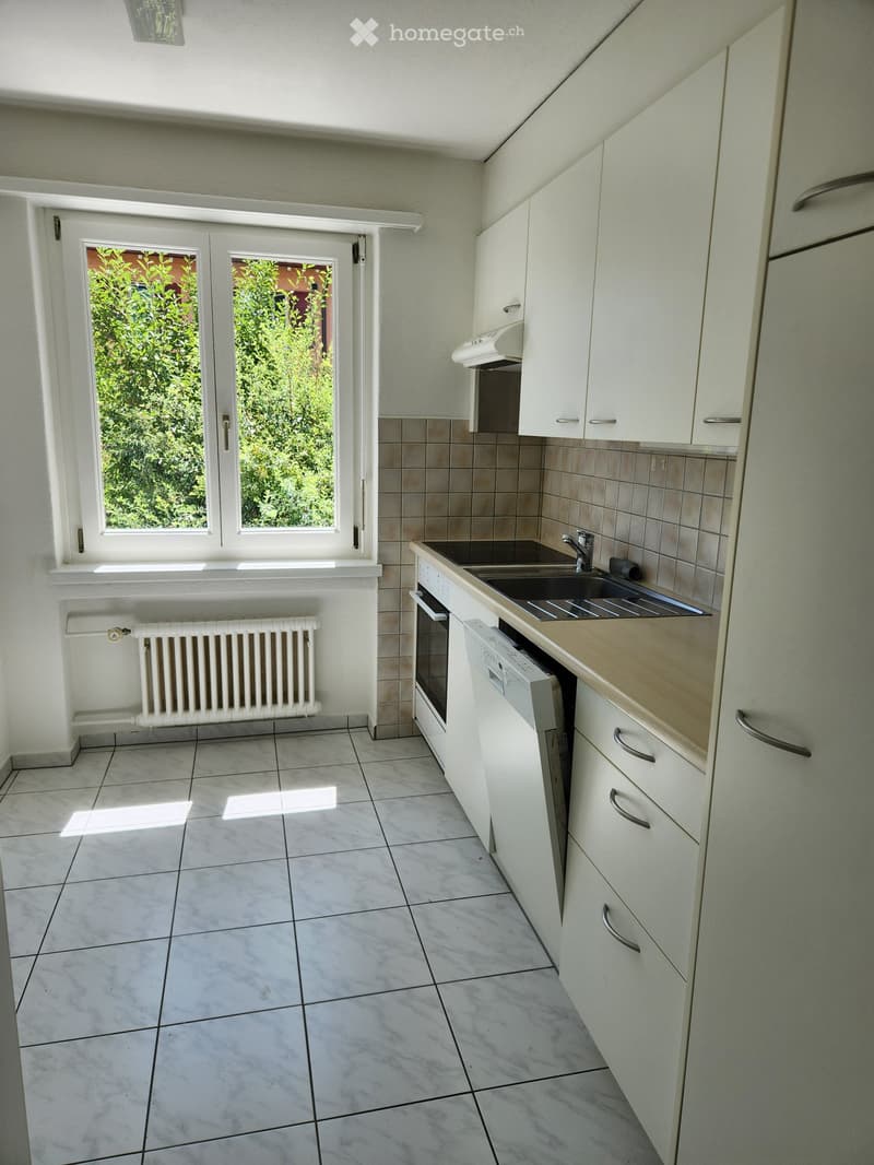 Modern kitchen with white cabinets, tiled floor, sink, oven, and dishwasher. Large window with view of greenery.