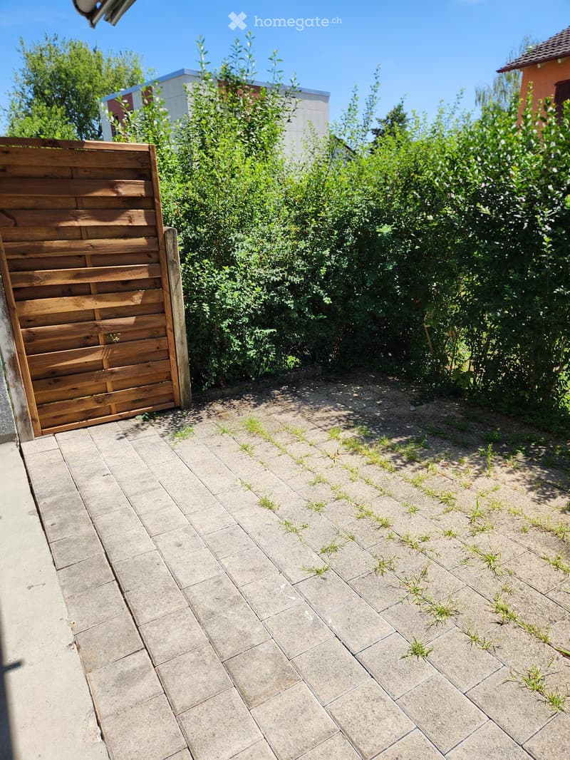 A paved outdoor area with a wooden gate, surrounded by greenery and neighboring buildings.