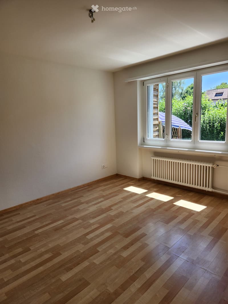 Empty room with wooden flooring, white walls, large windows, radiator, and visible sunlight patches on the floor.