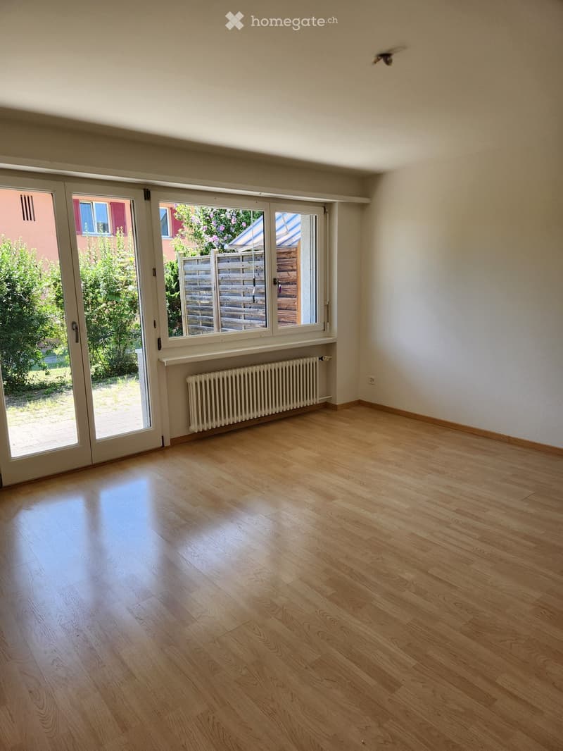 Empty room with light wooden flooring, white walls, large sliding glass doors leading to a garden, and a radiator below the windows.