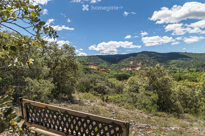 A scenic outdoor view from a wooden bench, overlooking a lush, green valley with trees, hills, and a partly cloudy blue sky.