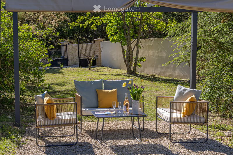 A serene outdoor seating area with a sofa, two armchairs, a coffee table, and decorative pillows, surrounded by greenery and a gravel ground.