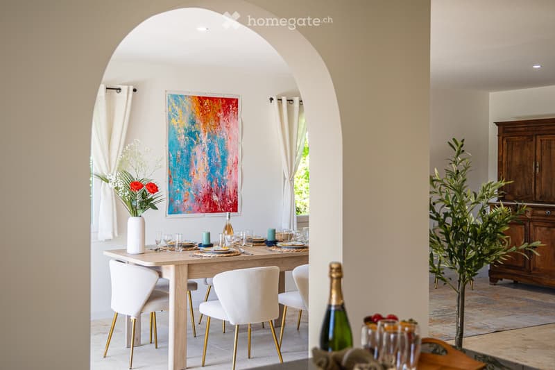Elegant dining room with a wooden table, white chairs with gold legs, a vase with flowers, and a colorful abstract painting on the wall.