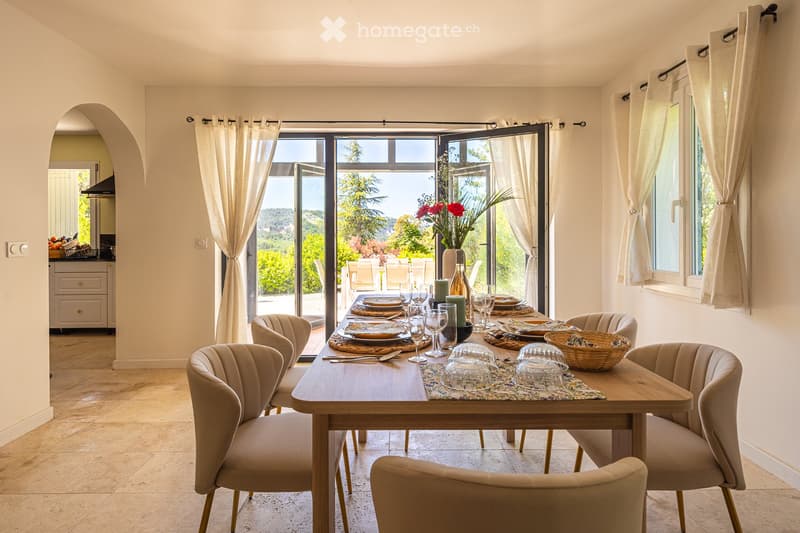 Elegant dining room with a wooden dining table set for a meal, surrounded by six upholstered chairs. Large windows with white curtains offer a scenic view of the garden. A vase with red flowers and a wine bottle are placed on the table.