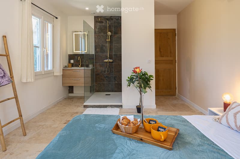 Cozy bedroom with a large bed, a tray with pastries and cups, a plant, a modern bathroom with a glass shower enclosure, and a wooden door leading to another room.