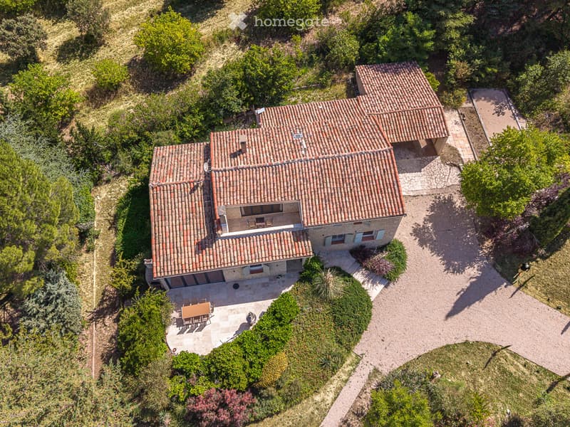 Aerial view of a two-story house with terracotta tile roof, surrounded by greenery and a gravel driveway. Features include a patio with outdoor furniture and a small balcony on the upper level.