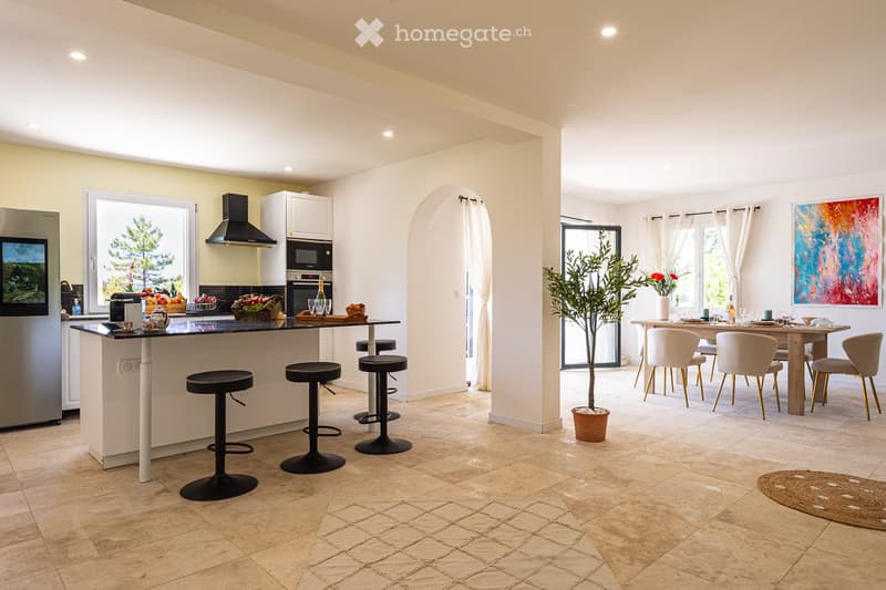 Open-plan kitchen and dining area with modern appliances, bar stools, dining table, chairs, and a decorative painting on the wall.