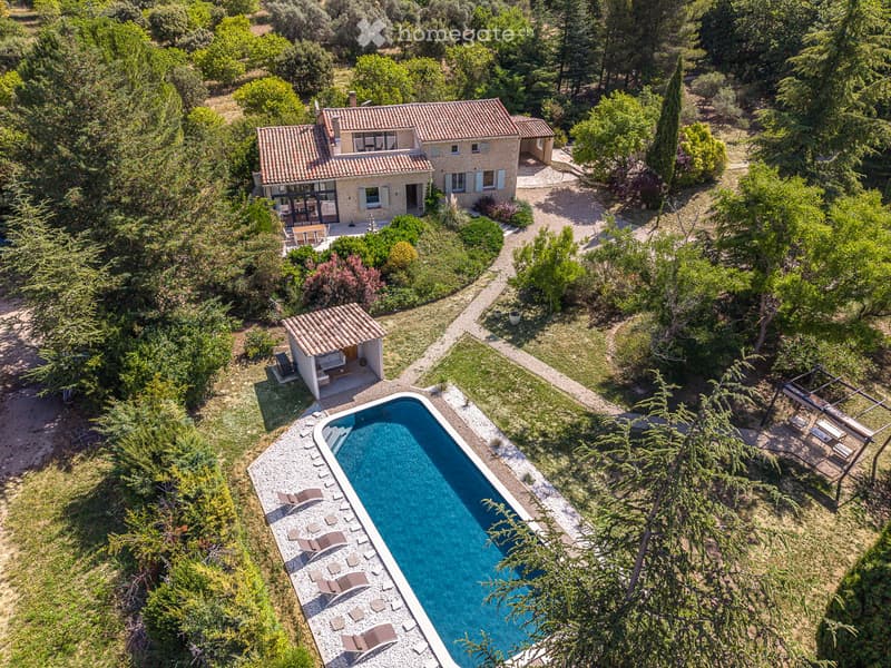 Aerial view of a house with a swimming pool, surrounded by greenery and trees. The pool is rectangular with lounge chairs around it, and there is a smaller structure nearby.