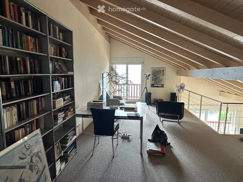 A well-lit attic room with a slanted wooden ceiling, featuring a large bookshelf filled with books, a desk with a chair, a printer, and a fan. There is a tripod near a window with a view, and a small seating area with a chair and a plant.