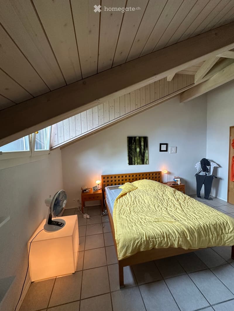 Cozy attic bedroom with slanted wooden ceiling, large window, bed with yellow quilt, nightstands with lamps, fan, and artwork on wall.