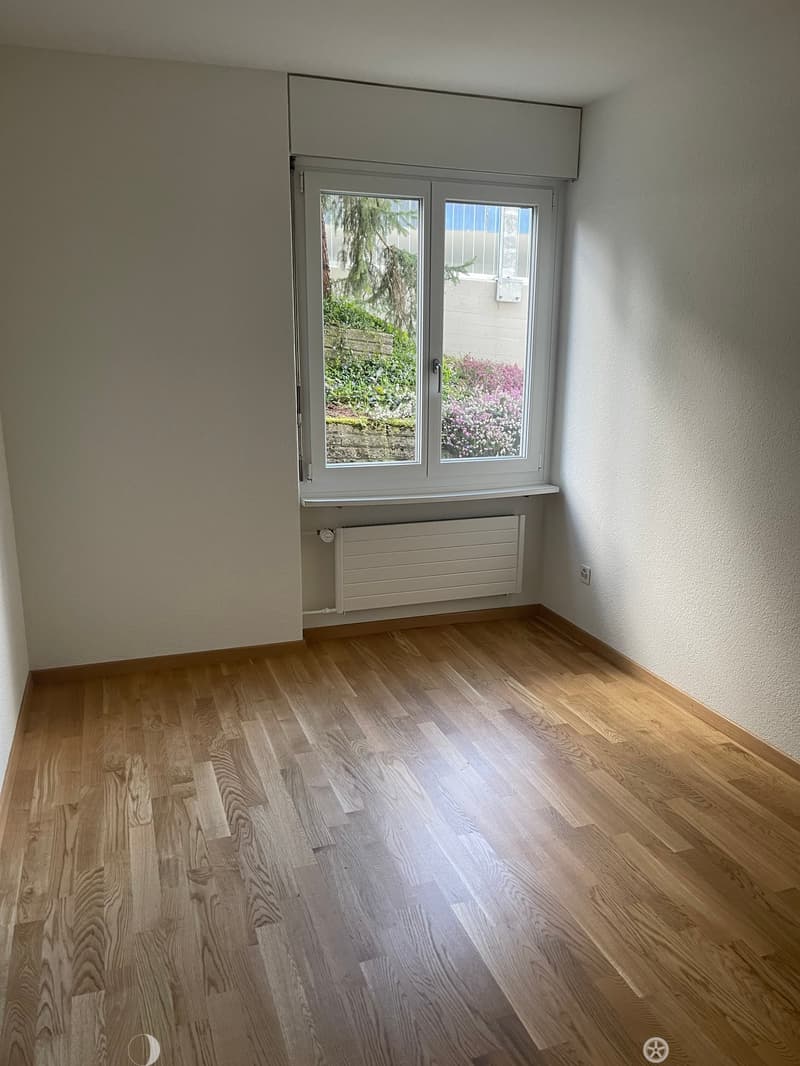 Empty room with wooden flooring, white walls, a window with a view of a garden, and a radiator below the window.