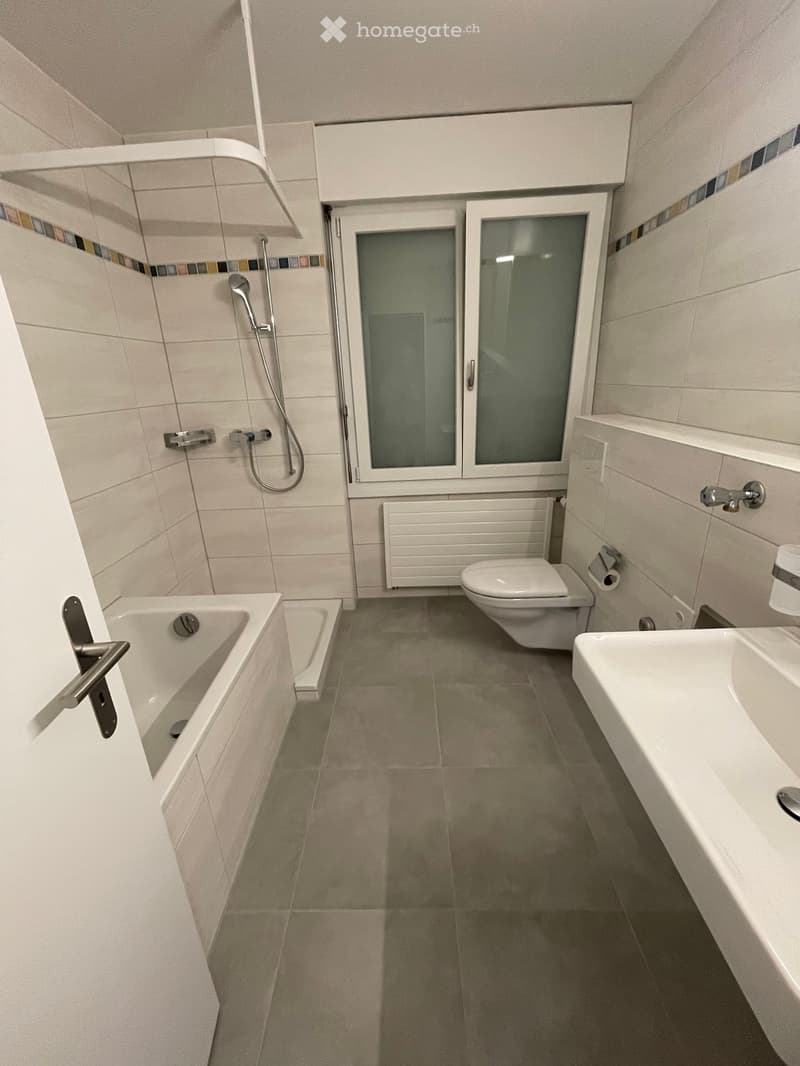 Modern bathroom with a bathtub, toilet, sink, shower, and radiator. Features tiled walls and floor, and a window with frosted glass panels.