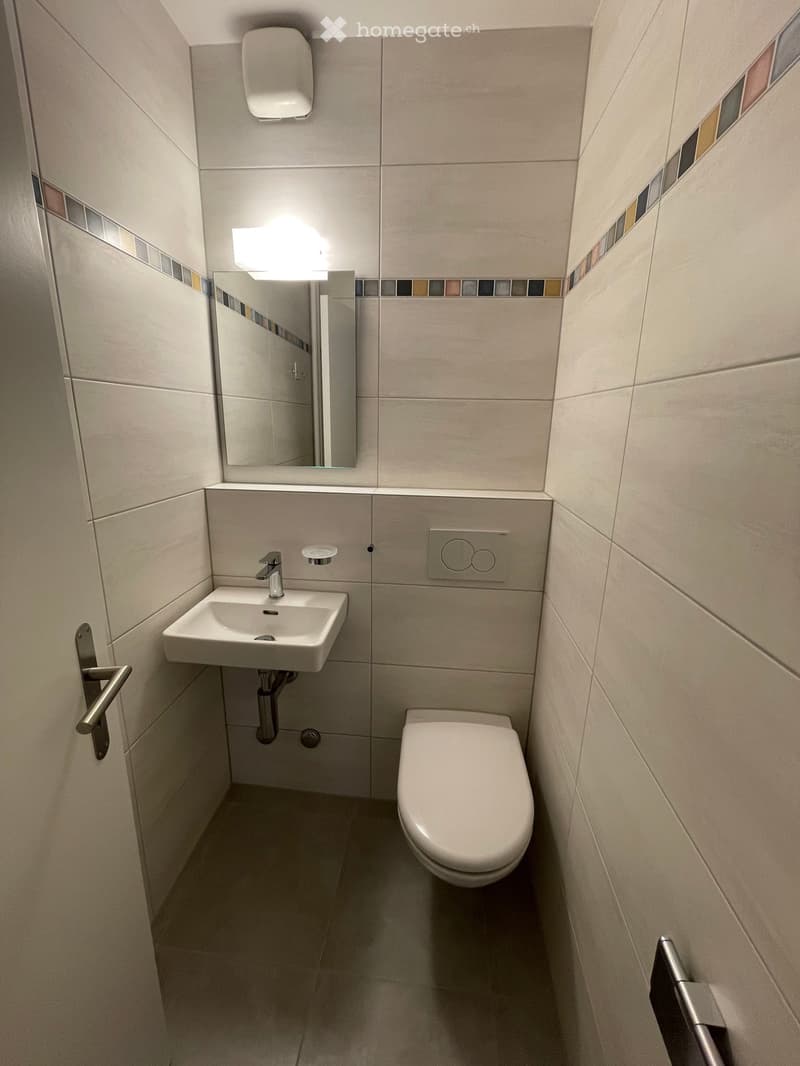 Modern bathroom with a white sink, chrome faucet, toilet, and a mirror above the sink. The walls are tiled with a decorative border, and the floor is tiled.