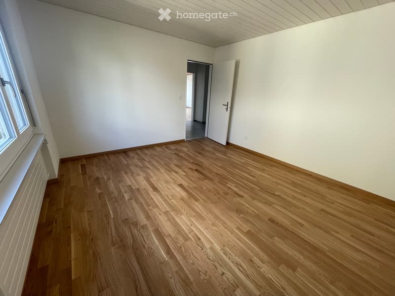 Empty room with light wooden flooring, white walls, and a window on one side. A door is open to an adjacent space.