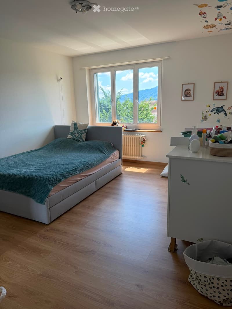 Bright bedroom with wooden flooring, large windows offering a scenic view, a grey sofa with a teal blanket, a small table with various items, and a framed picture on the wall.