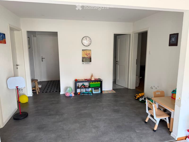 A well-lit, open-plan room with grey tiled flooring, white walls, and several doors leading to other areas. The room contains a small table with chairs, a ball, a shelf with toys, a stool, and a clock on the wall.