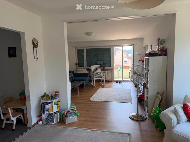 A well-lit living room with wooden flooring, a sofa, a coffee table, a fan, a bookshelf, and various toys and decorations.