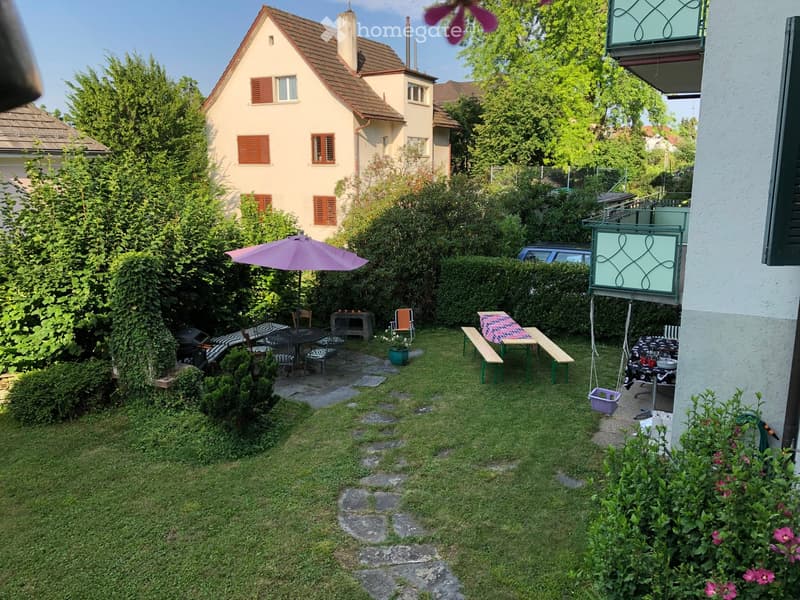 Garden with lawn, patio area, umbrella, tables, chairs, benches, and a small table with a pink tablecloth.