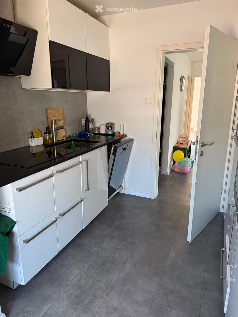 Modern kitchen with white cabinets, black countertops, stainless steel sink, dishwasher, and various small appliances. A hallway leads to another room with toys visible.