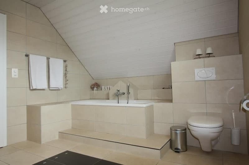 Modern bathroom with a bathtub, toilet, towels, and a trash can. The walls are tiled in beige, and there is a small decorative sign that reads "BATH."