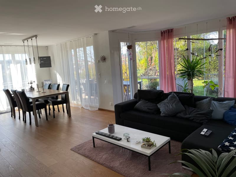 Modern living and dining area with a black sectional sofa, white coffee table, dining table with chairs, and large windows with sheer curtains. The space features wooden flooring and a cozy rug.