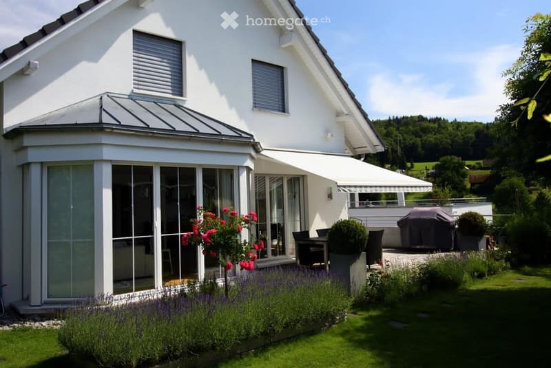 Modern two-story house with white exterior, large windows, covered patio, and well-maintained garden with flowers and shrubs.