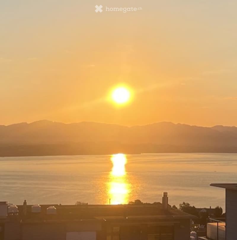 Balcony or terrace view overlooking a lake at sunset, with mountains in the background and the sun reflecting on the water.