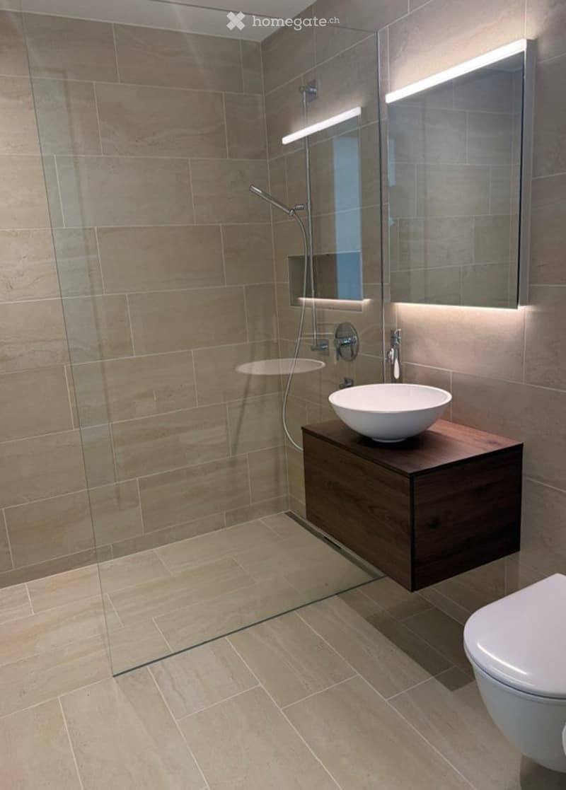 Modern bathroom with a glass-enclosed shower, a floating wooden sink cabinet, a round white sink, and a toilet. The walls and floor are tiled in a light beige color, and the shower area features built-in lighting.
