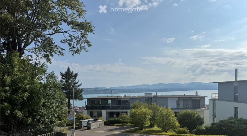 Modern two-story house with large windows and balconies, situated on a hillside overlooking a body of water and distant mountains.