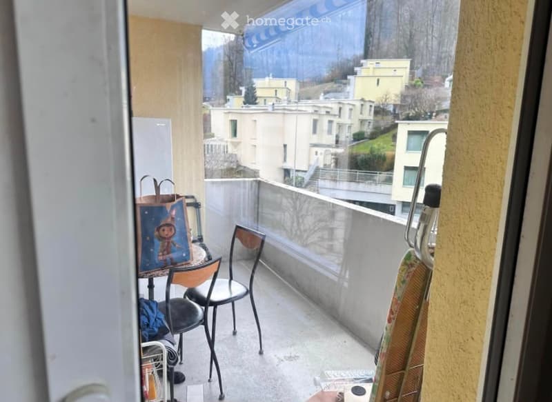 Balcony with a view of the city and surrounding buildings, featuring a small table, chairs, and a few personal items.