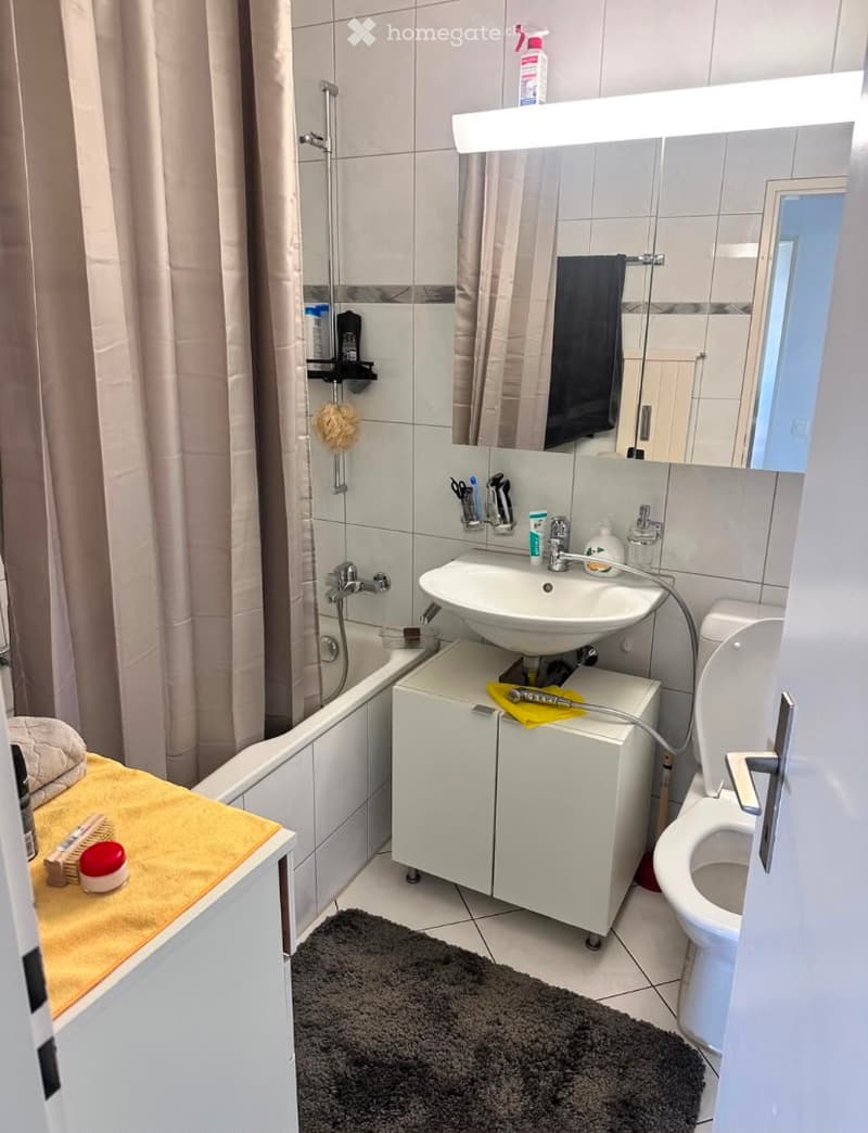 Modern bathroom with a bathtub, sink, toilet, and a large mirror. Includes a white cabinet under the sink, a towel on the side, and cleaning supplies on the shelf.