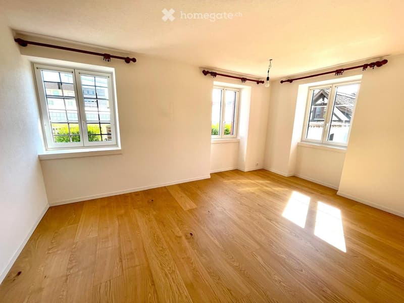 Empty room with light wooden flooring and white walls, featuring three windows with white frames and a red curtain rod above them.