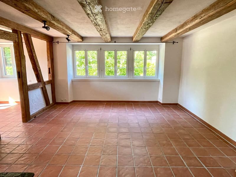 Empty room with exposed wooden beams, tiled floor, and large windows with white frames.