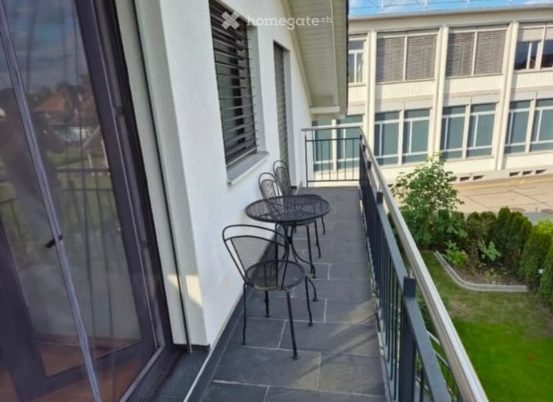 Balcony with a small table and chairs, overlooking a landscaped area with greenery and a building in the background.