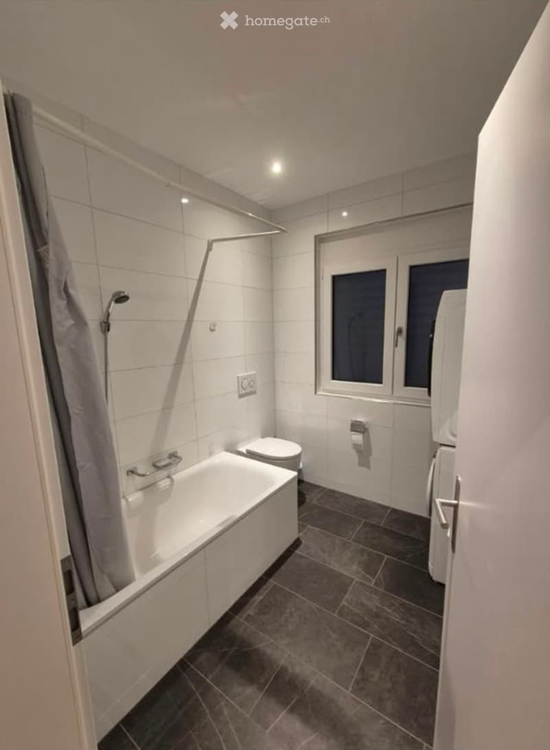 Modern bathroom with a white bathtub, toilet, shower, and a towel hanging on the wall. The room features white tiled walls and dark grey tiled flooring.