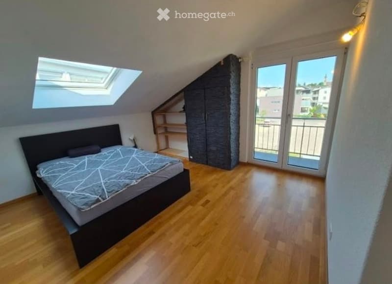Spacious attic bedroom with wooden flooring, large window, skylight, and access to a balcony. Features a dark wooden bed, wardrobe, and built-in shelves.