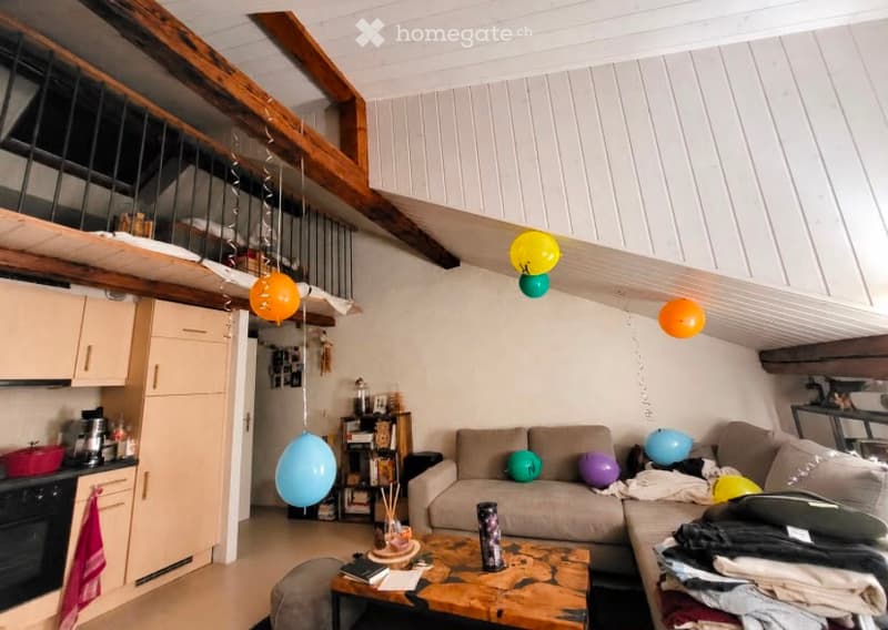Living room with exposed wooden beams, a grey sectional sofa, a wooden coffee table, a bookshelf, and a small kitchen area with an oven. Several colorful balloons are floating in the air.