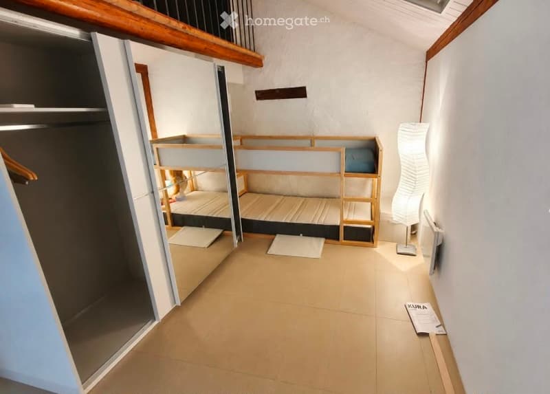 A compact room with a wooden bunk bed, a wardrobe, and a modern floor lamp. The room features light-colored tiled flooring and white walls.