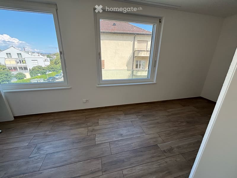 Empty room with wooden flooring and large windows offering a view of the street and neighboring buildings.