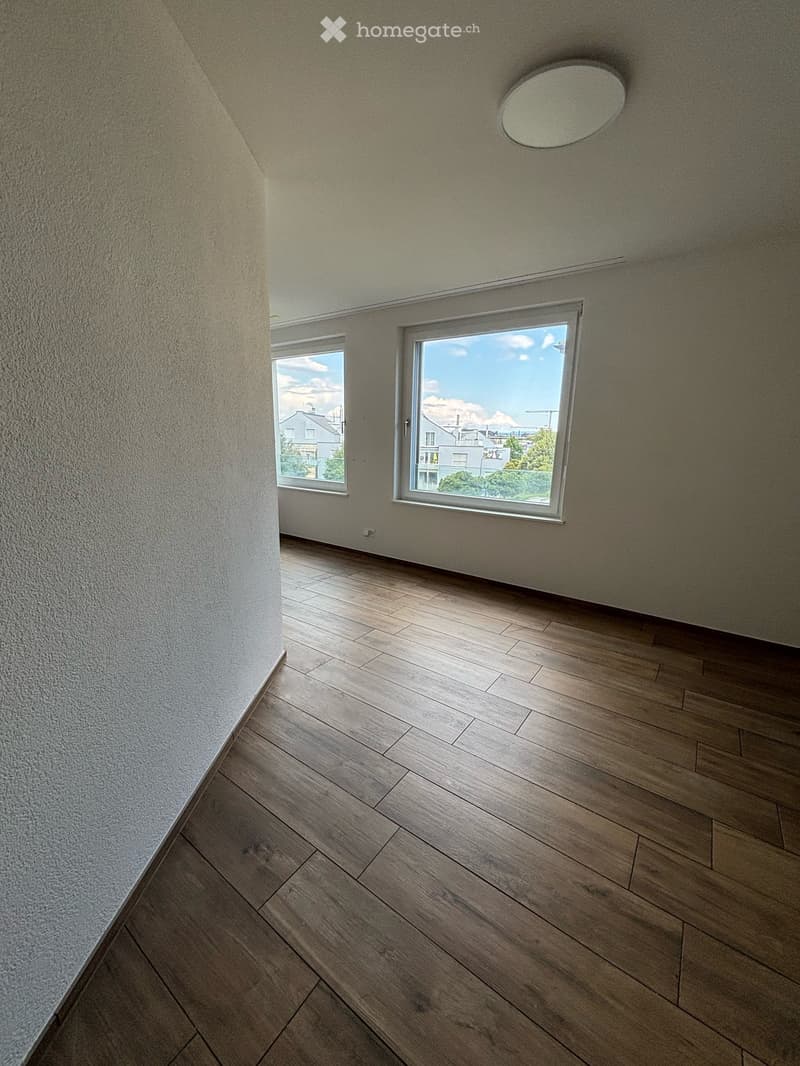 Empty room with wooden flooring, white walls, and large windows offering a view of the outside. The ceiling features a round light fixture.
