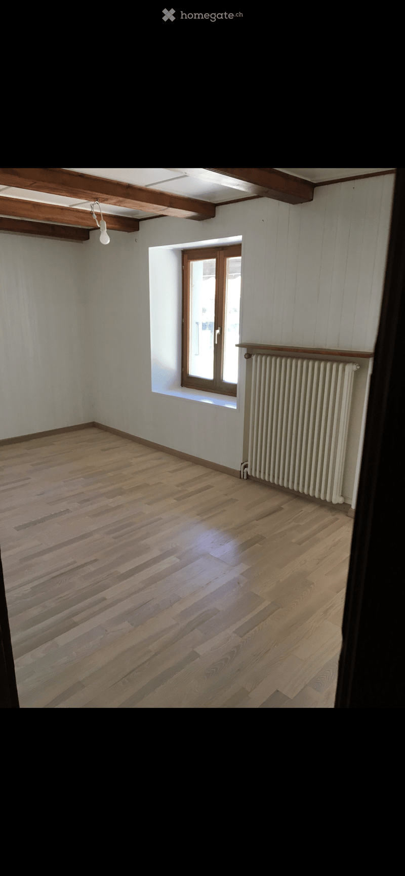 Empty room with wooden flooring, white walls, exposed wooden beams on the ceiling, a radiator, and a window with a wooden frame.